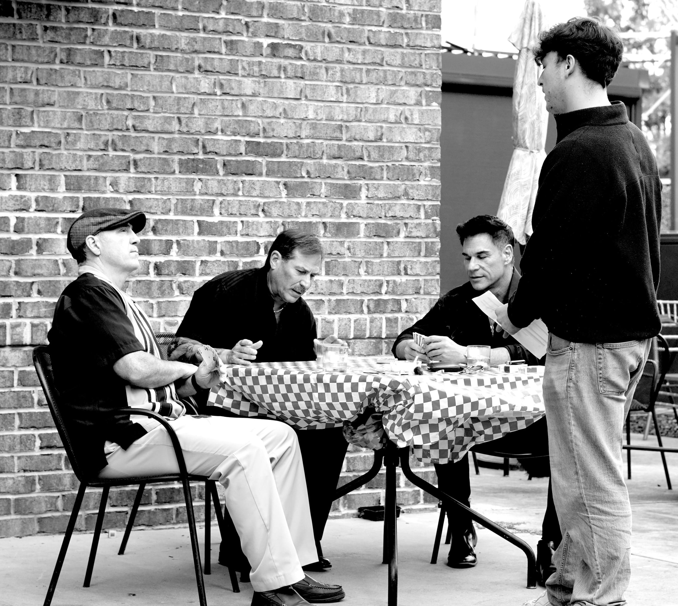 Three men playing cards at a checkered table while a fourth stands over them